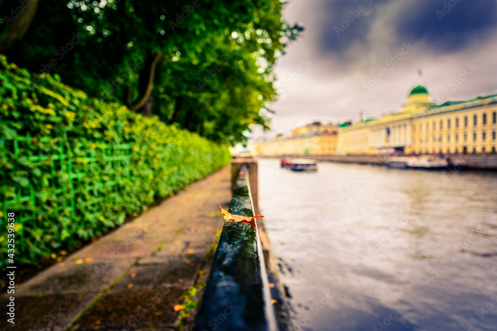 Rainy autumn day in the city, alley in the park near the river on which the ship is sailing. Close up view from the handrail on which lies the oak leaf on the embankment level Rainy autumn day in the city, alley in the park near the river on which the ship is sailing. Close up view from the handrail on which lies the oak leaf on the embankment level