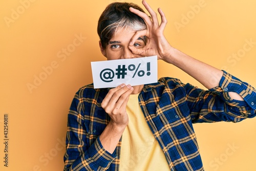 Young hispanic man covering mouth with insult message paper smiling happy doing ok sign with hand on eye looking through fingers