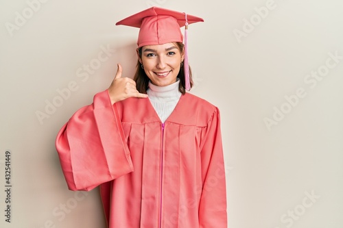 Young caucasian woman wearing graduation cap and ceremony robe smiling doing phone gesture with hand and fingers like talking on the telephone. communicating concepts.