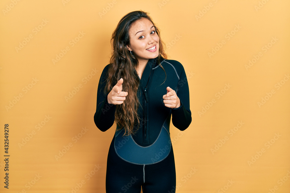 Young hispanic girl wearing diver neoprene uniform pointing fingers to camera with happy and funny face. good energy and vibes. Young hispanic girl wearing diver neoprene uniform pointing fingers to camera with happy and funny face. good energy and vibes.