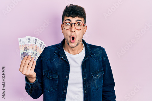 Young hispanic man holding icelandic krona banknotes scared and amazed with open mouth for surprise, disbelief face photo