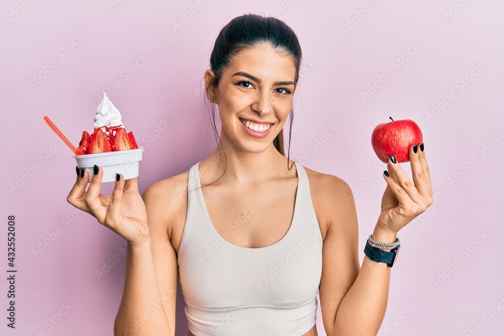 Young hispanic woman wearing sportswear holding apple and ice cream smiling with a happy and cool smile on face. showing teeth. Young hispanic woman wearing sportswear holding apple and ice cream smiling with a happy and cool smile on face. showing teeth.