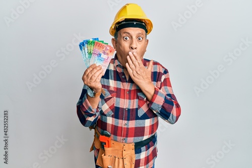 Senior hispanic man wearing handyman uniform and hardhat covering mouth with hand, shocked and afraid for mistake. surprised expression