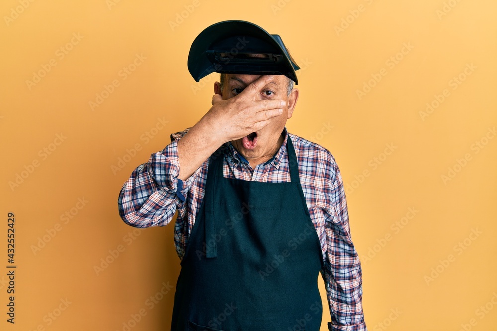 Handsome senior man with grey hair wearing welding protection mask peeking in shock covering face and eyes with hand, looking through fingers with embarrassed expression. Handsome senior man with grey hair wearing welding protection mask peeking in shock covering face and eyes with hand, looking through fingers with embarrassed expression.