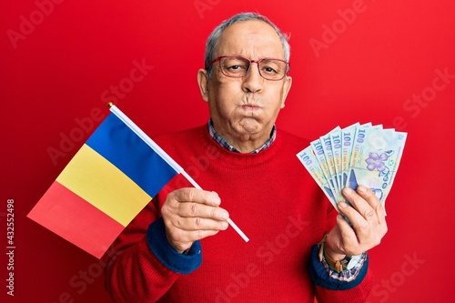 Handsome senior man with grey hair holding romania flag and leu banknotes puffing cheeks with funny face. mouth inflated with air, catching air.