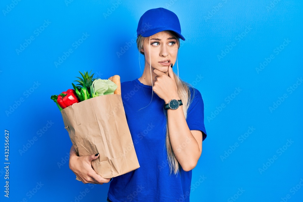 Young caucasian woman wearing courier uniform with groceries from supermarket thinking worried about a question, concerned and nervous with hand on chin Young caucasian woman wearing courier uniform with groceries from supermarket thinking worried about a question, concerned and nervous with hand on chin