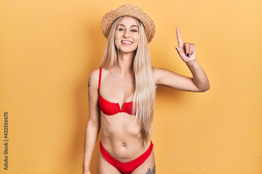 Young caucasian woman wearing bikini and summer hat showing and pointing up with finger number one while smiling confident and happy. Young caucasian woman wearing bikini and summer hat showing and pointing up with finger number one while smiling confident and happy.
