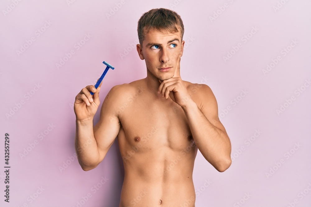 Fototapeta premium Young caucasian man shirtless holding razor serious face thinking about question with hand on chin, thoughtful about confusing idea