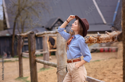 a girl with cowboy clothes with a hat stands on a horse farm near the fence