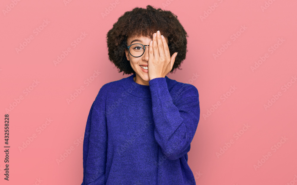 Young hispanic girl wearing casual winter sweater and glasses covering one eye with hand, confident smile on face and surprise emotion. Young hispanic girl wearing casual winter sweater and glasses covering one eye with hand, confident smile on face and surprise emotion.