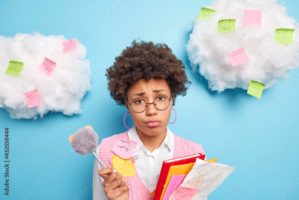 Indoor shot of frustrated unhappy Afro American schoolgirl going to do home assignment holds folders with papers and pen wears round spectacles neat clothes makes notes on stickers pasted around Indoor shot of frustrated unhappy Afro American schoolgirl going to do home assignment holds folders with papers and pen wears round spectacles neat clothes makes notes on stickers pasted around