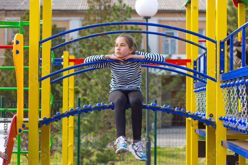 Brunette girl 10 years old, bored on the playground, sits on a slide with her legs dangling. 