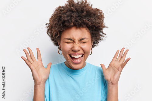 Overjoyed ethnic woman with curly bushy hair reacts emotionally on unexpected gift smiles broadly closes eyes wears blue t shirt isolated over white background. People emotions reactions concept