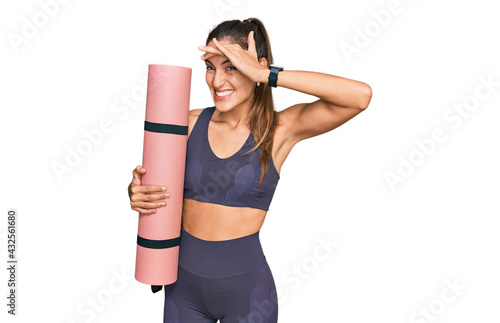 Young beautiful woman wearing sportswear holding yoga mat stressed and frustrated with hand on head, surprised and angry face