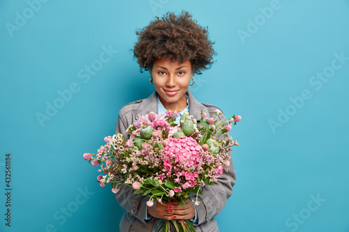 Portrait of satisfied young African American woman holds nice bouquet of flowers looks confidently at camera wears grey jacket isolated over blue background has pleasant appearance comes on date