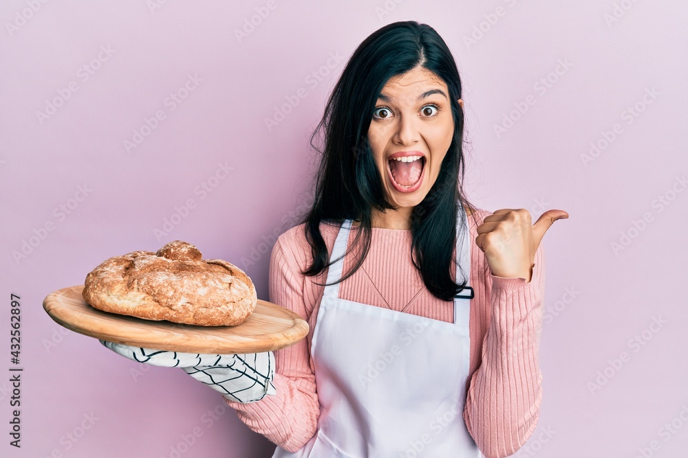 Young hispanic woman wearing baker uniform holding homemade bread pointing thumb up to the side smiling happy with open mouth Young hispanic woman wearing baker uniform holding homemade bread pointing thumb up to the side smiling happy with open mouth