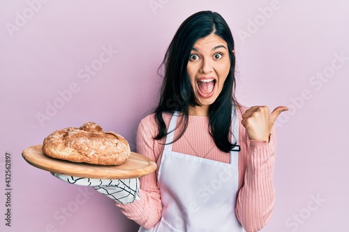 Young hispanic woman wearing baker uniform holding homemade bread pointing thumb up to the side smiling happy with open mouth