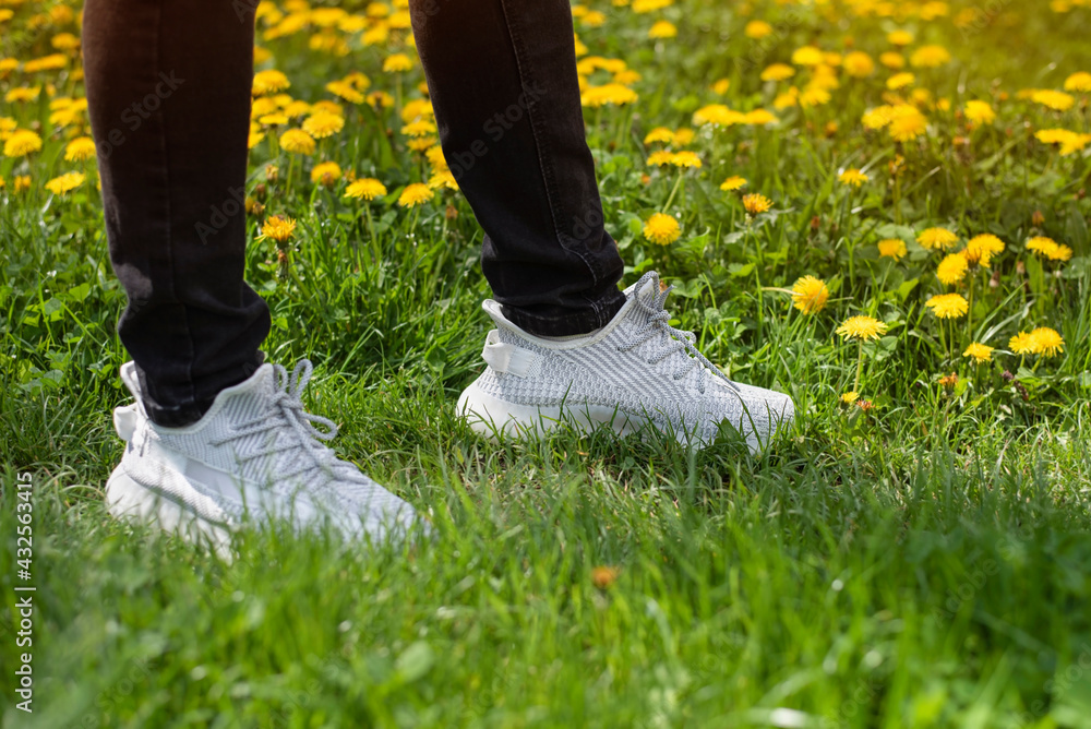 Among the green grass and yellow dandelions blooming in the spring is a guy in gray sneakers made of fabric, the focus on sneakers