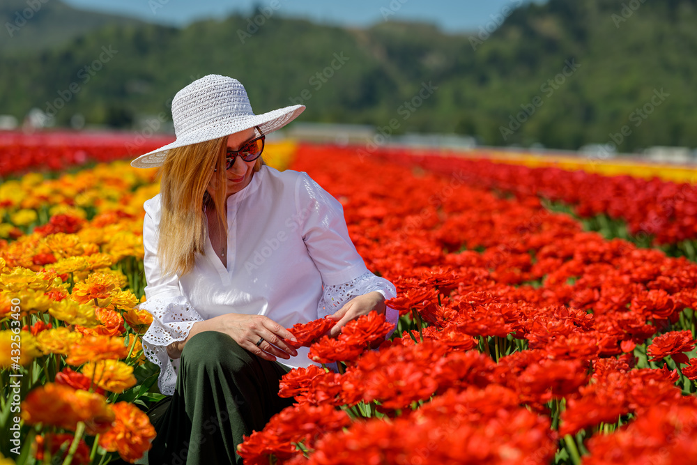 Well dressed middle aged woman wearing white straw hat, sitting between the rows of an amazing tulip plantation, showing love and enjoying the view of the vibrant tulips in the field