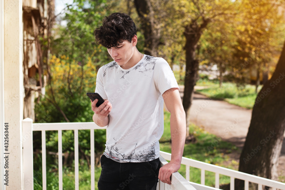 A teenage boy is waiting for his girlfriend near the house and looks at the phone, reads the news. The guy has a serious look with a phone in his hand