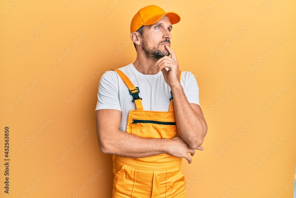 Young handsome man wearing handyman uniform over yellow background with hand on chin thinking about question, pensive expression. smiling with thoughtful face. doubt concept. Young handsome man wearing handyman uniform over yellow background with hand on chin thinking about question, pensive expression. smiling with thoughtful face. doubt concept.