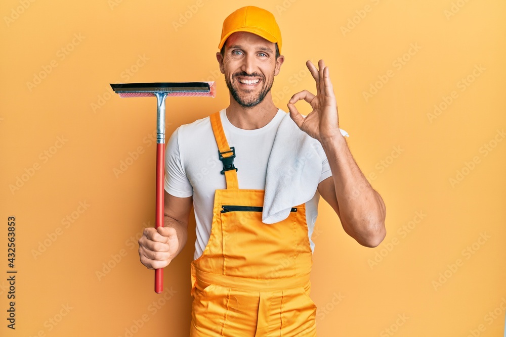 Young handsome man wearing glass clenaer uniform and squeegee doing ok sign with fingers, smiling friendly gesturing excellent symbol Young handsome man wearing glass clenaer uniform and squeegee doing ok sign with fingers, smiling friendly gesturing excellent symbol