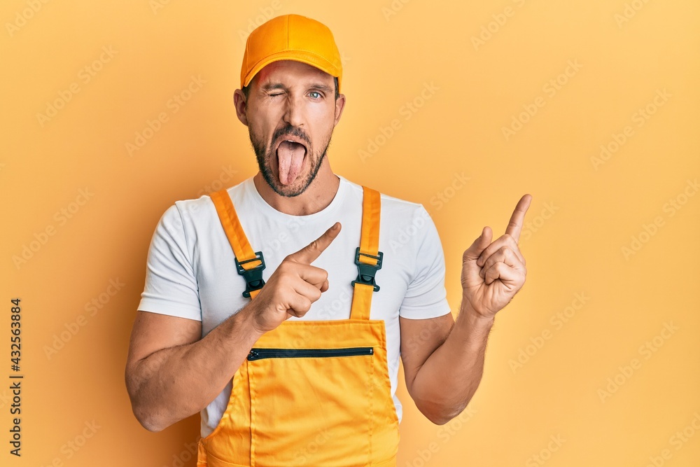 Young handsome man wearing handyman uniform pointing to the side sticking tongue out happy with funny expression. Young handsome man wearing handyman uniform pointing to the side sticking tongue out happy with funny expression.