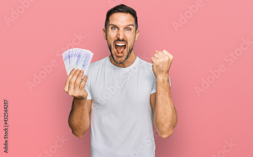 Handsome man with beard holding 20 swedish krona banknotes screaming proud, celebrating victory and success very excited with raised arms © Krakenimages.com