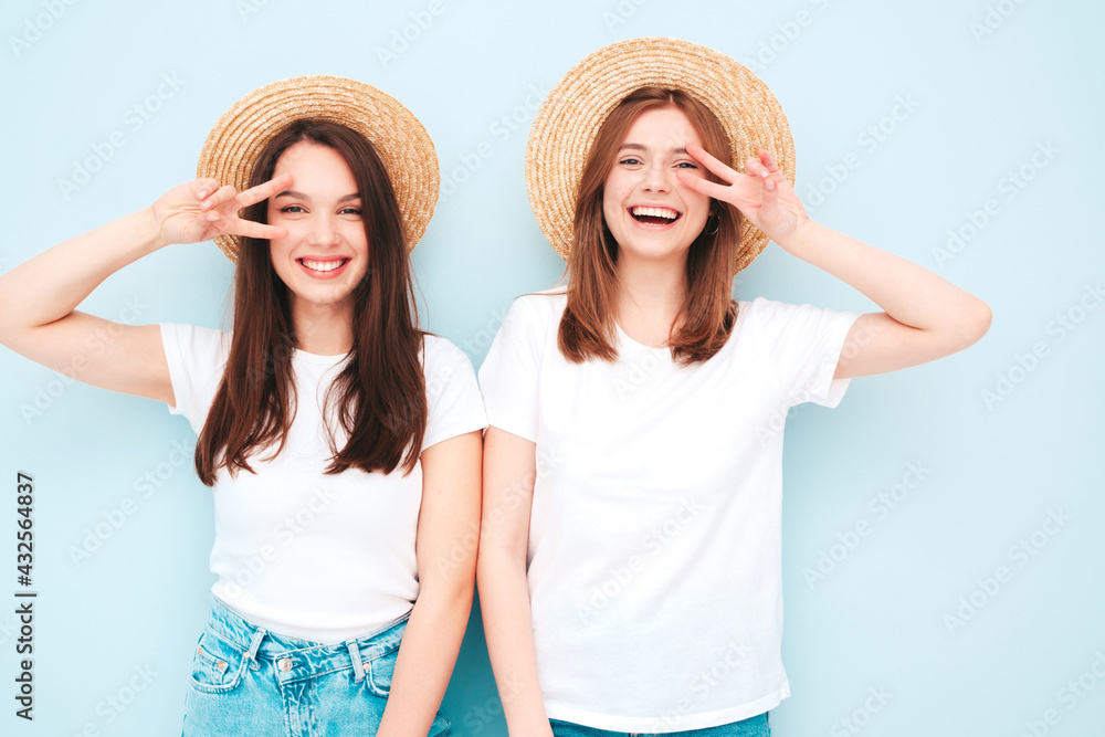 Two young beautiful smiling hipster female in trendy summer white t-shirt and jeans clothes.Sexy carefree women posing near light blue wall in studio.Positive models in hats in sunglasses