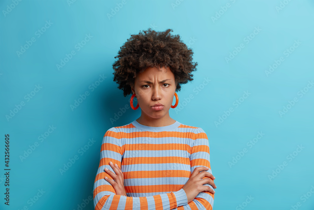 Sulking displeased young African American being angry and insulted about unfair situation purses lips looks disappointed keeps arms folded wears striped jumper isolated over blue background. Sulking displeased young African American being angry and insulted about unfair situation purses lips looks disappointed keeps arms folded wears striped jumper isolated over blue background.