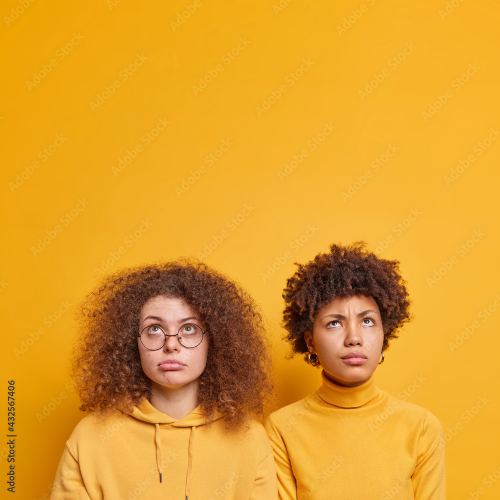 Two unhappy curly haired women of different nationalities focused above on something upleasant stand shoulder to shoulder isolated over yellow background with copy space for your advertising content.