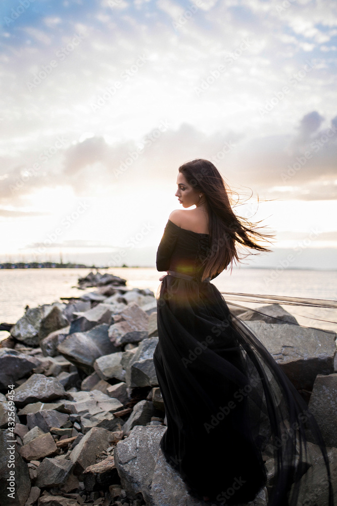 beautiful sensual woman in black long dress posing on the rocks by the sea during sunset. beautiful sensual woman in black long dress posing on the rocks by the sea during sunset.