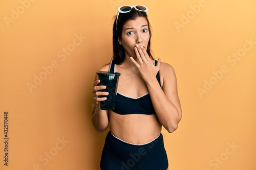 Young latin woman wearing bikini drinking soda covering mouth with hand, shocked and afraid for mistake. surprised expression