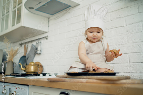 Little blonde chef girl is playing in the kitchen with cookie and dessert molds. A healthy child and a cheerful child's mood. Bright kitchen in the apartment.