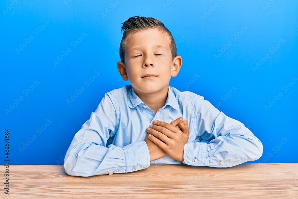 Adorable caucasian kid wearing casual clothes sitting on the table smiling with hands on chest, eyes closed with grateful gesture on face. health concept. Adorable caucasian kid wearing casual clothes sitting on the table smiling with hands on chest, eyes closed with grateful gesture on face. health concept.