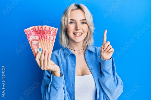 Young blonde girl holding philippine peso banknotes smiling with an idea or question pointing finger with happy face  number one