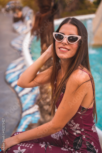 A young brunette in a maroon floral dress and trendy white sunglasses enjoying the summer in the golden hour at a city fountain © unai