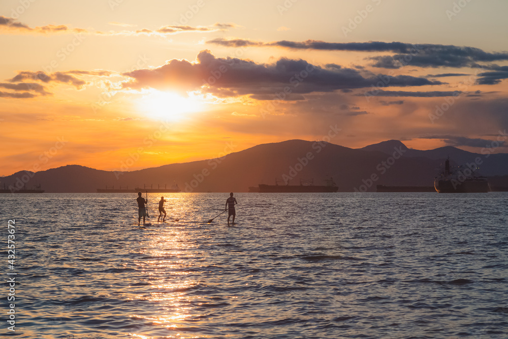 Fototapeta premium Silhouette of paddle boarders during sunset golden hour light on on open Pacific ocean water at Kitsilano Beach on an idyllic summer evening in Vancouver, B.C., Canada.