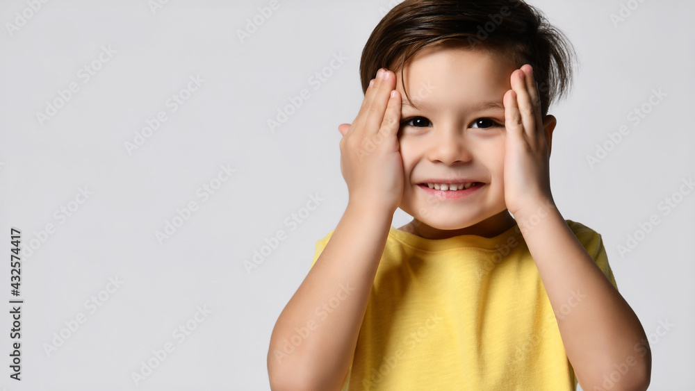 Studio shot of an emotionally adorable little boy raising his eyebrows and covering his temples with his hands, surprised and shocked, showing a true astonished reaction to unexpected news