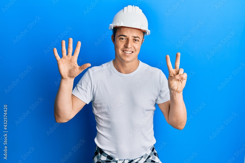 Handsome young man wearing builder uniform and hardhat showing and pointing up with fingers number seven while smiling confident and happy.