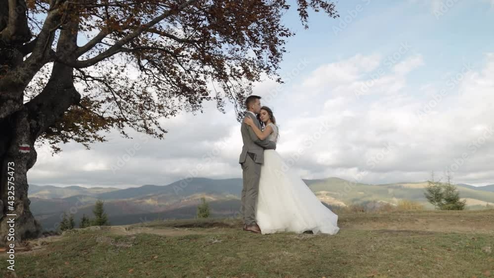 Newlyweds. Lovely caucasian bride and groom on cloudy sky background. Embracing, hugging. Happy wedding couple family on mountain autumn slope hill. Man woman in love. Bride in gorgeous wedding dress Newlyweds. Lovely caucasian bride and groom on cloudy sky background. Embracing, hugging. Happy wedding couple family on mountain autumn slope hill. Man woman in love. Bride in gorgeous wedding dress