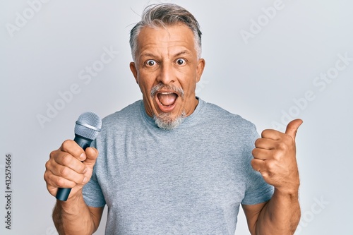 Middle age grey-haired man singing song using microphone pointing thumb up to the side smiling happy with open mouth
