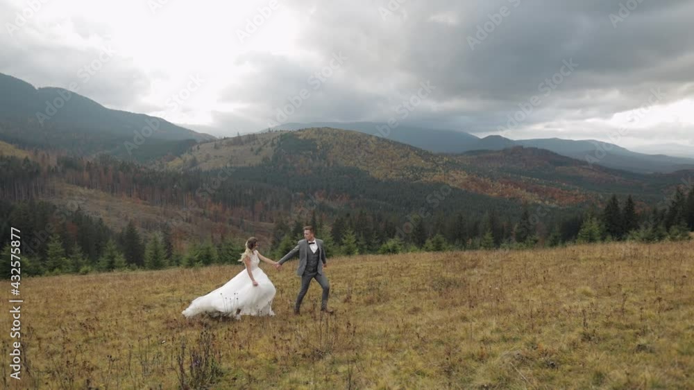Newlyweds. Lovely young caucasian bride and groom running, holding hands on mountain slope hill. Wedding couple. Happy family. Man woman in love. Autumn. Bride in gorgeous wedding dress. Slow motion Newlyweds. Lovely young caucasian bride and groom running, holding hands on mountain slope hill. Wedding couple. Happy family. Man woman in love. Autumn. Bride in gorgeous wedding dress. Slow motion