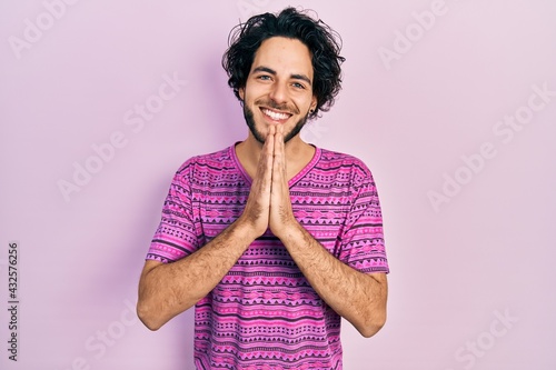 Handsome hispanic man wearing casual pink t shirt praying with hands together asking for forgiveness smiling confident.