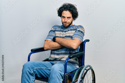 Handsome hispanic man sitting on wheelchair skeptic and nervous, disapproving expression on face with crossed arms. negative person.