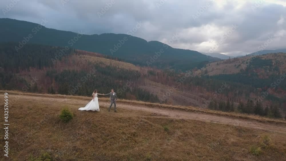 Aerial view. Lovely caucasian newlyweds bride and groom walking on mountain slope hill. Wedding couple making a kiss. Happy family of man woman in love. Bride in wedding dress. Cloudy sky background Aerial view. Lovely caucasian newlyweds bride and groom walking on mountain slope hill. Wedding couple making a kiss. Happy family of man woman in love. Bride in wedding dress. Cloudy sky background