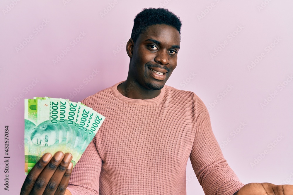 Handsome black man holding 20000 indonesian rupiah celebrating achievement with happy smile and winner expression with raised hand Handsome black man holding 20000 indonesian rupiah celebrating achievement with happy smile and winner expression with raised hand