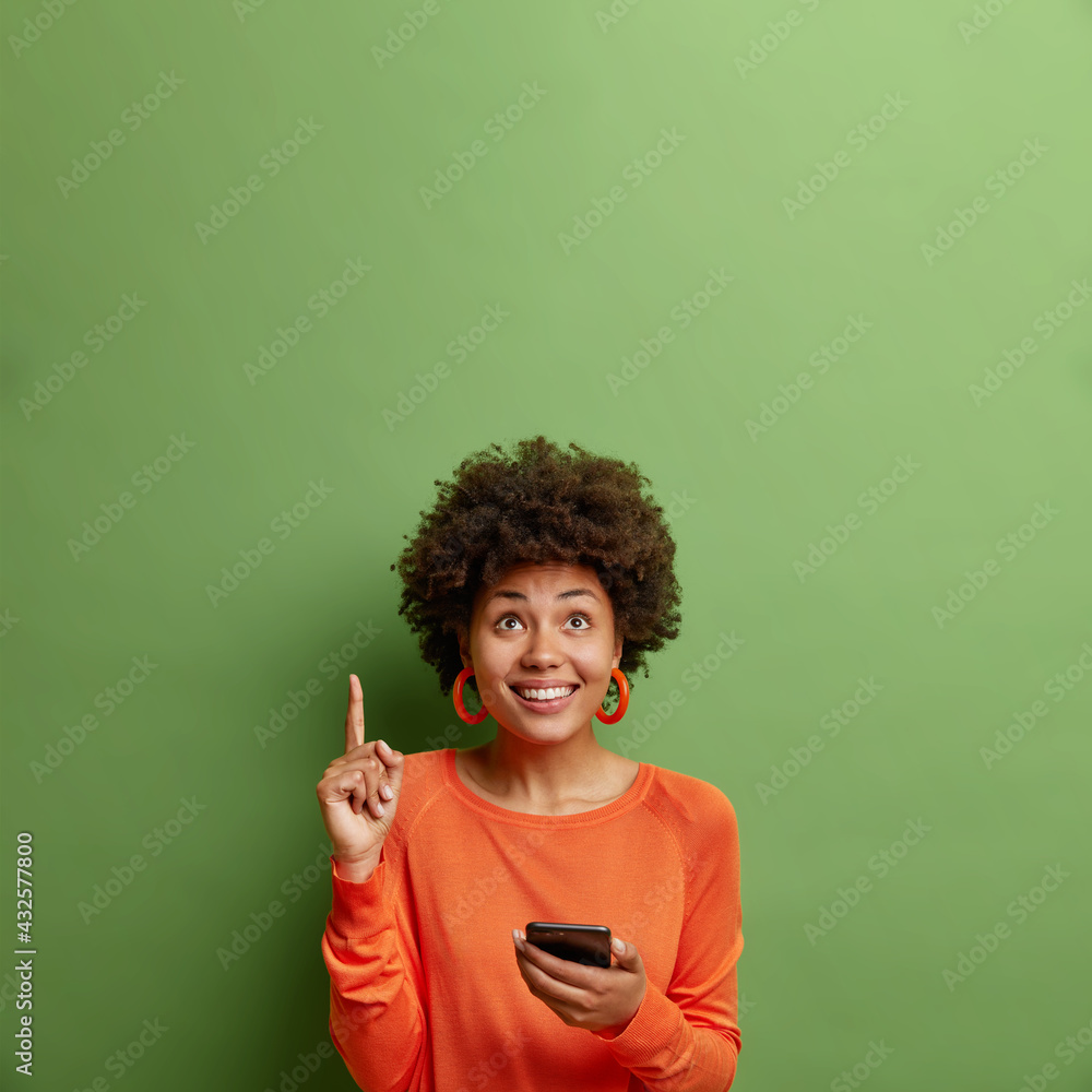 Vertical shot of positive young Afro American woman holds modern smartphone indicates upwards with index finger demonstrates copy space for your advertisement isolated over green background.