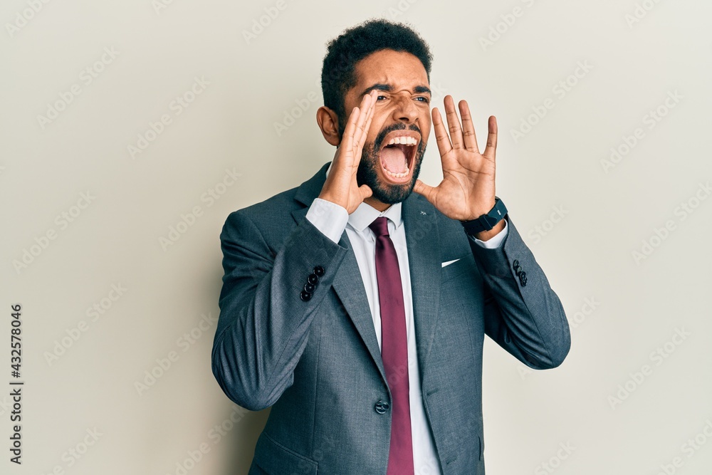 Handsome hispanic man with beard wearing business suit and tie shouting angry out loud with hands over mouth