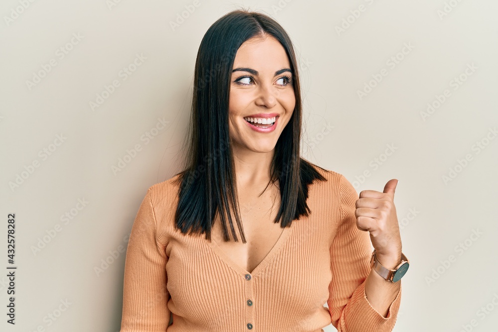 Young brunette woman wearing casual clothes pointing thumb up to the side smiling happy with open mouth Young brunette woman wearing casual clothes pointing thumb up to the side smiling happy with open mouth